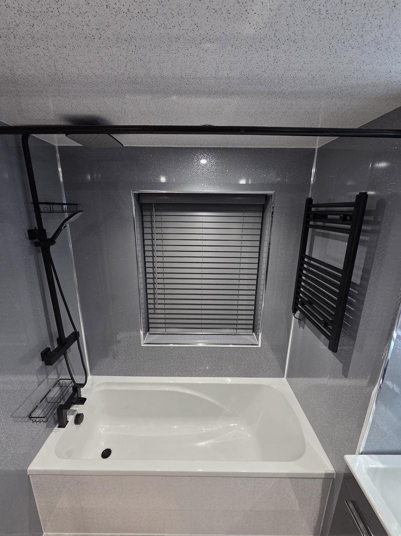 Bath with a black thermostatic shower riser and glass screen against grey wall panels