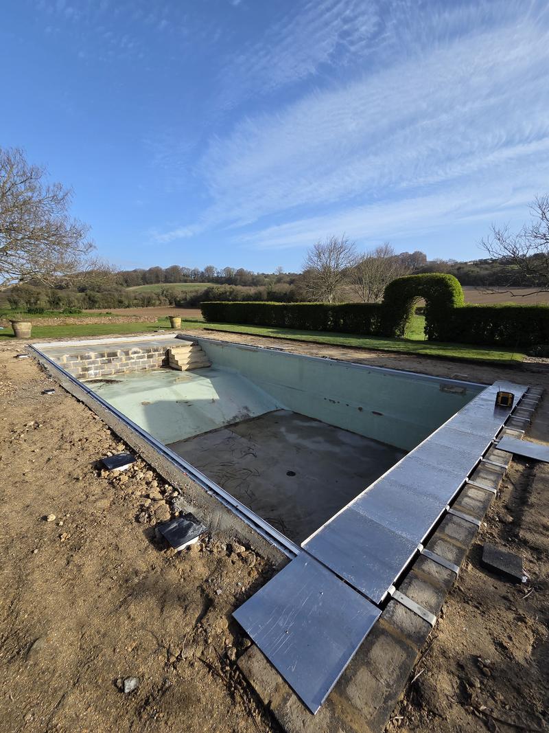 Drained swimming pool mid-refurbishment with new stone coping slabs being set out along both long edges