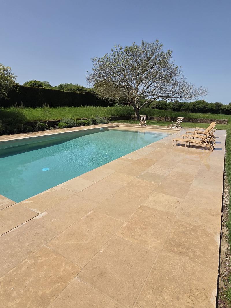 Finished swimming pool with a broad cream sandstone terrace, loungers along the far edge and mature planting behind