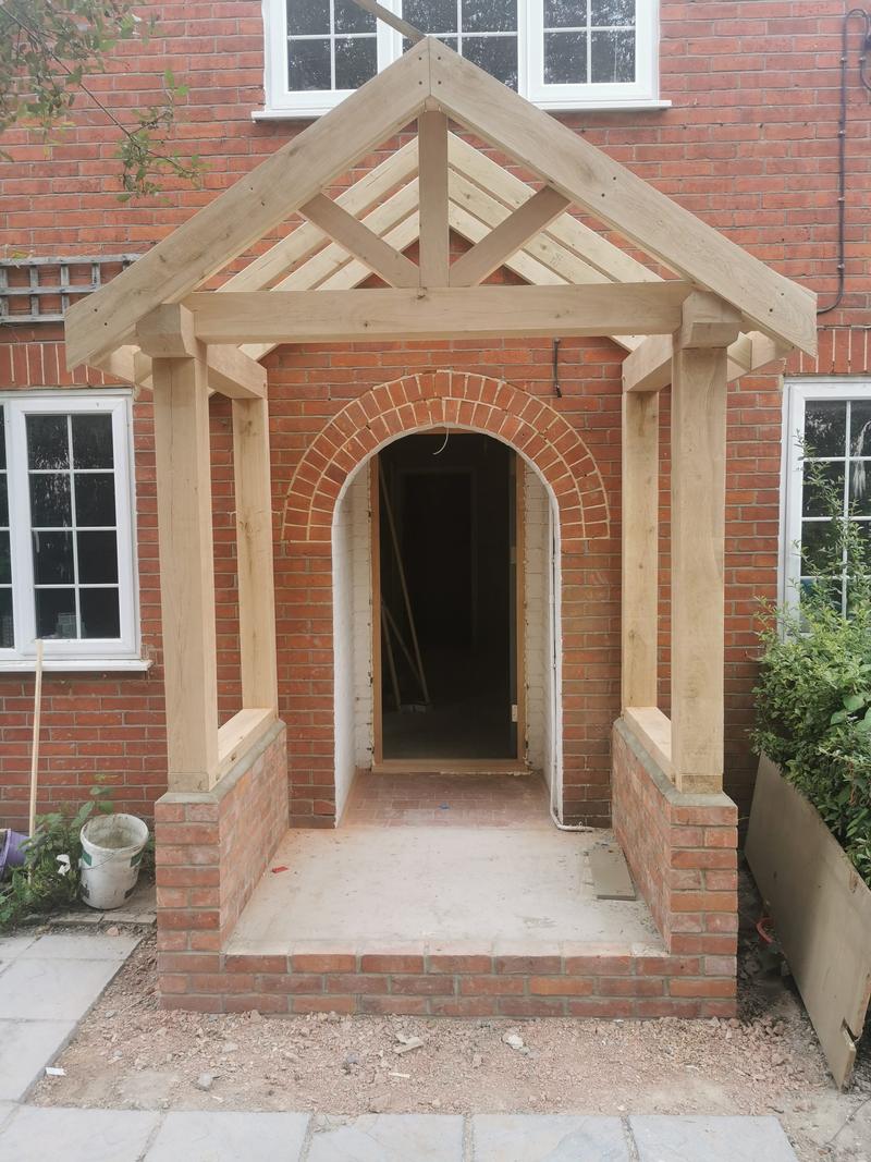 Green oak porch with a king post truss above a brick arched doorway, before the roof covering