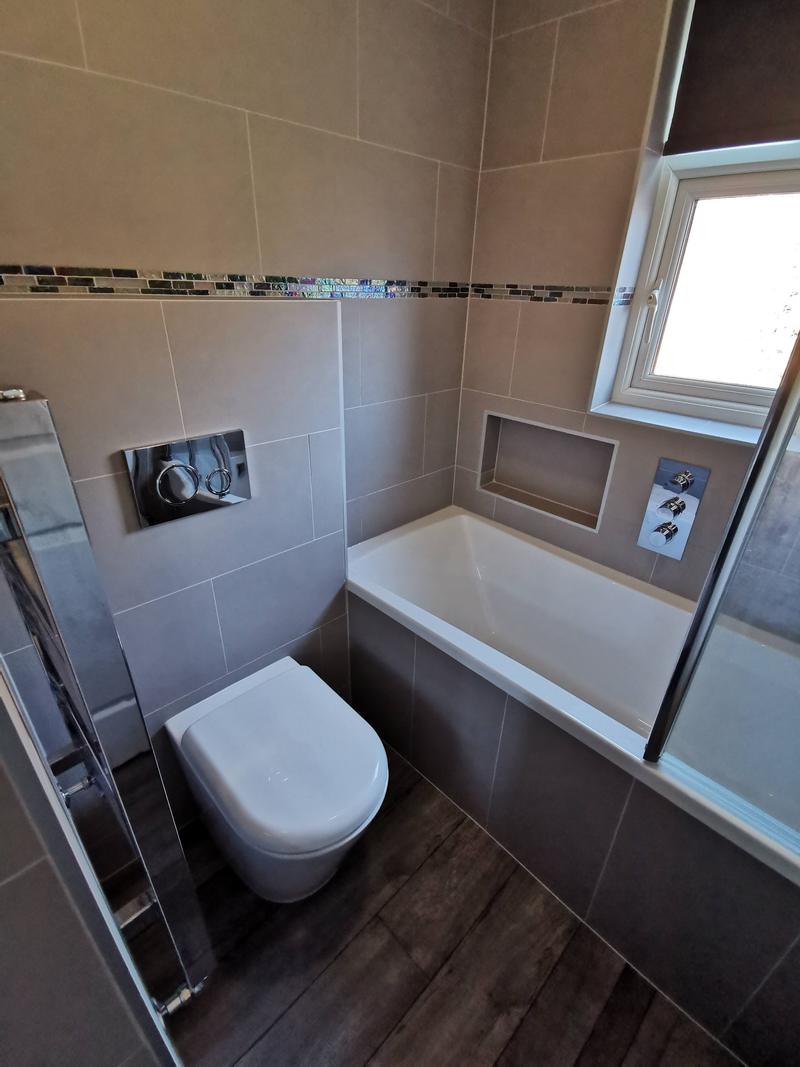 Grey tiled bathroom with a mosaic border, wall-hung WC and a bath with a shower screen