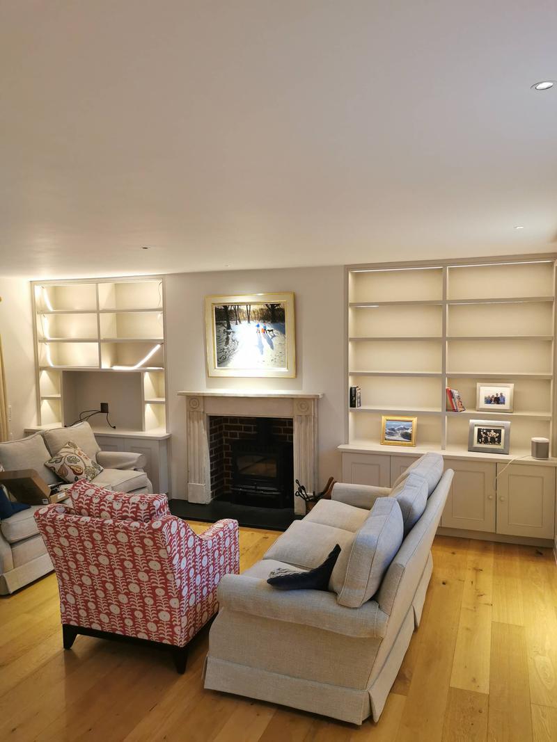 Sitting room with lit bespoke alcove bookcases either side of a stone fireplace and log burner